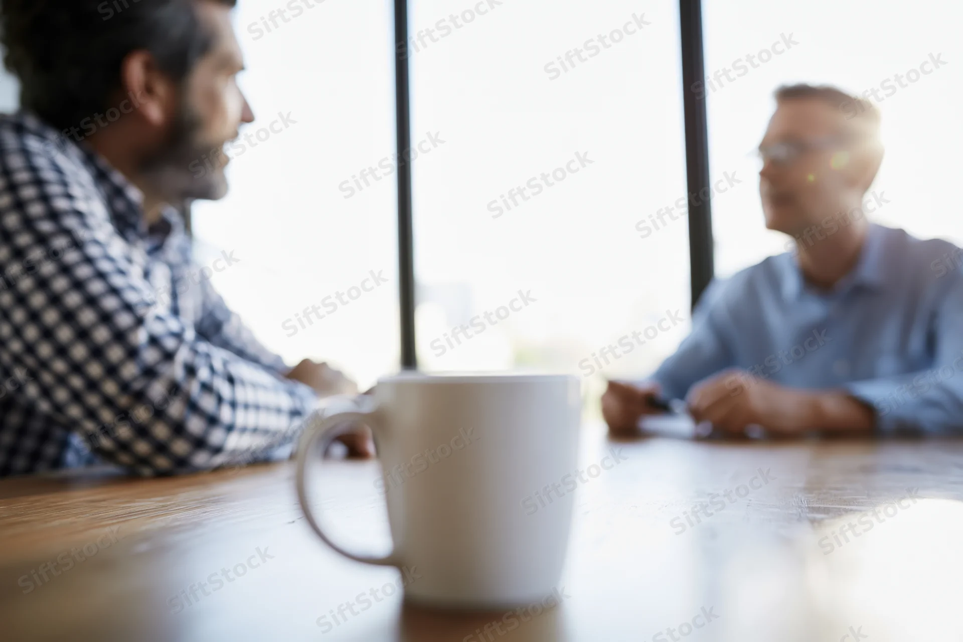 Two men in conversation across a wooden table in an office setting with a coffee mug in the foreground