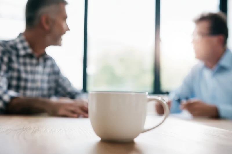 Two men in conversation across a wooden table in an office with a coffee cup in focus