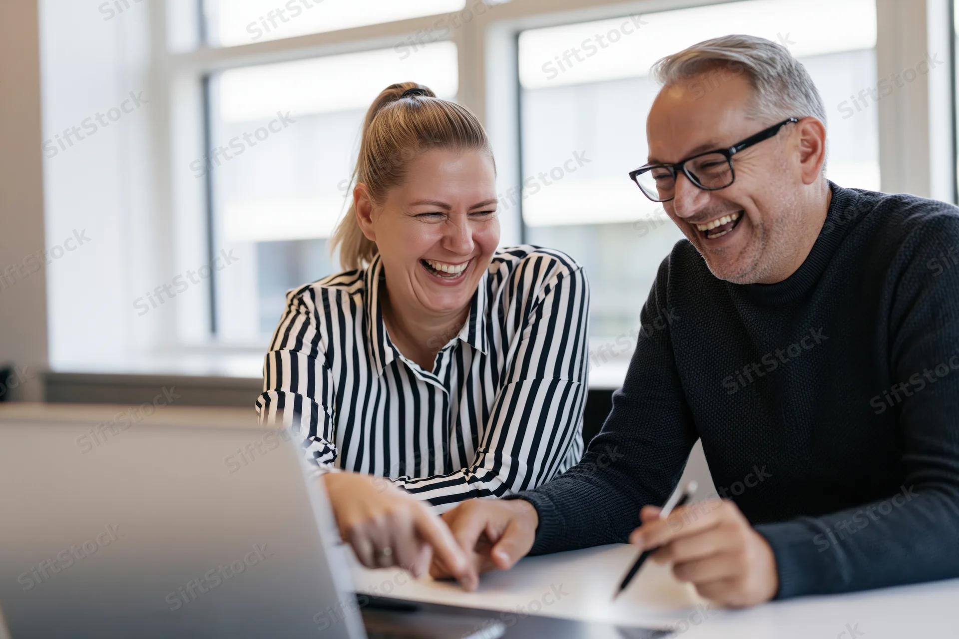 Two colleagues laughing while looking at a laptop screen in an office meeting