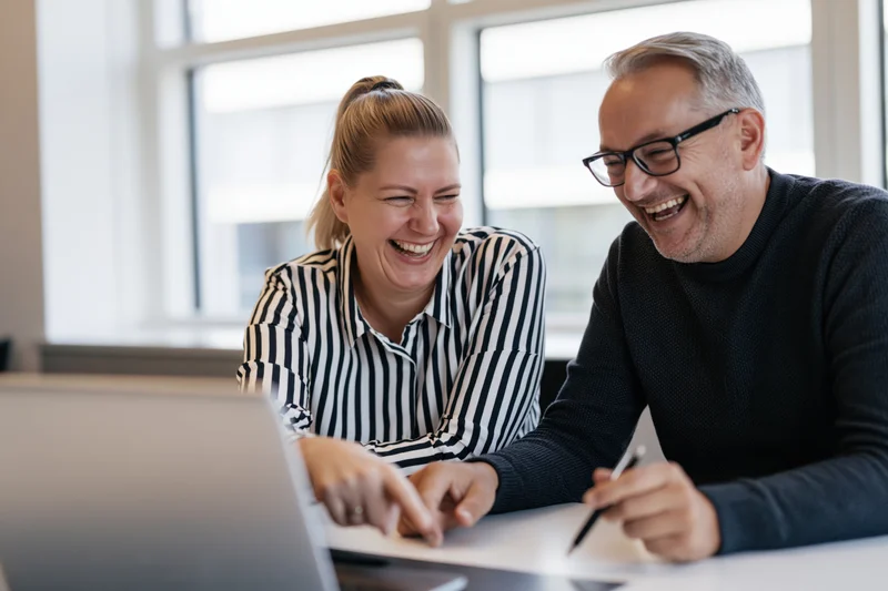 Two colleagues laughing while looking at a laptop screen in an office meeting