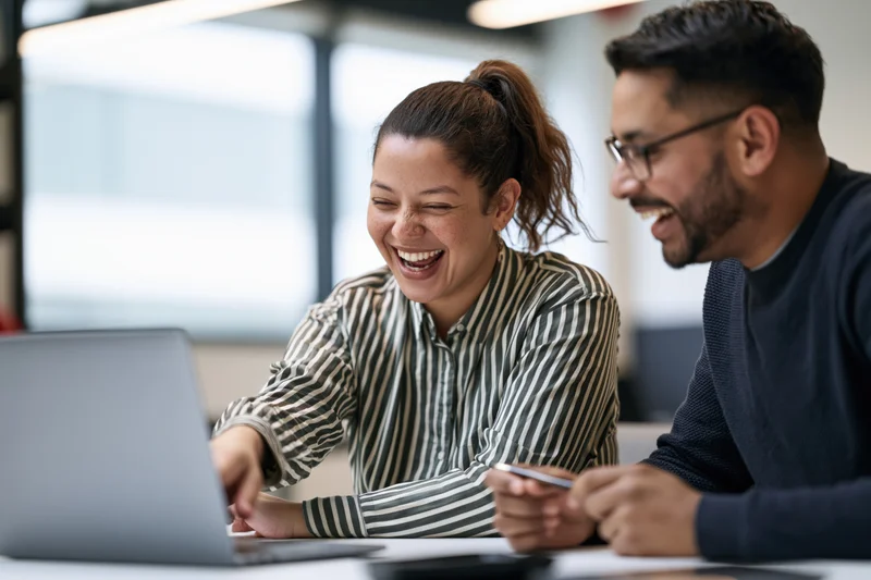 Two colleagues laughing while looking at a laptop screen in an office setting