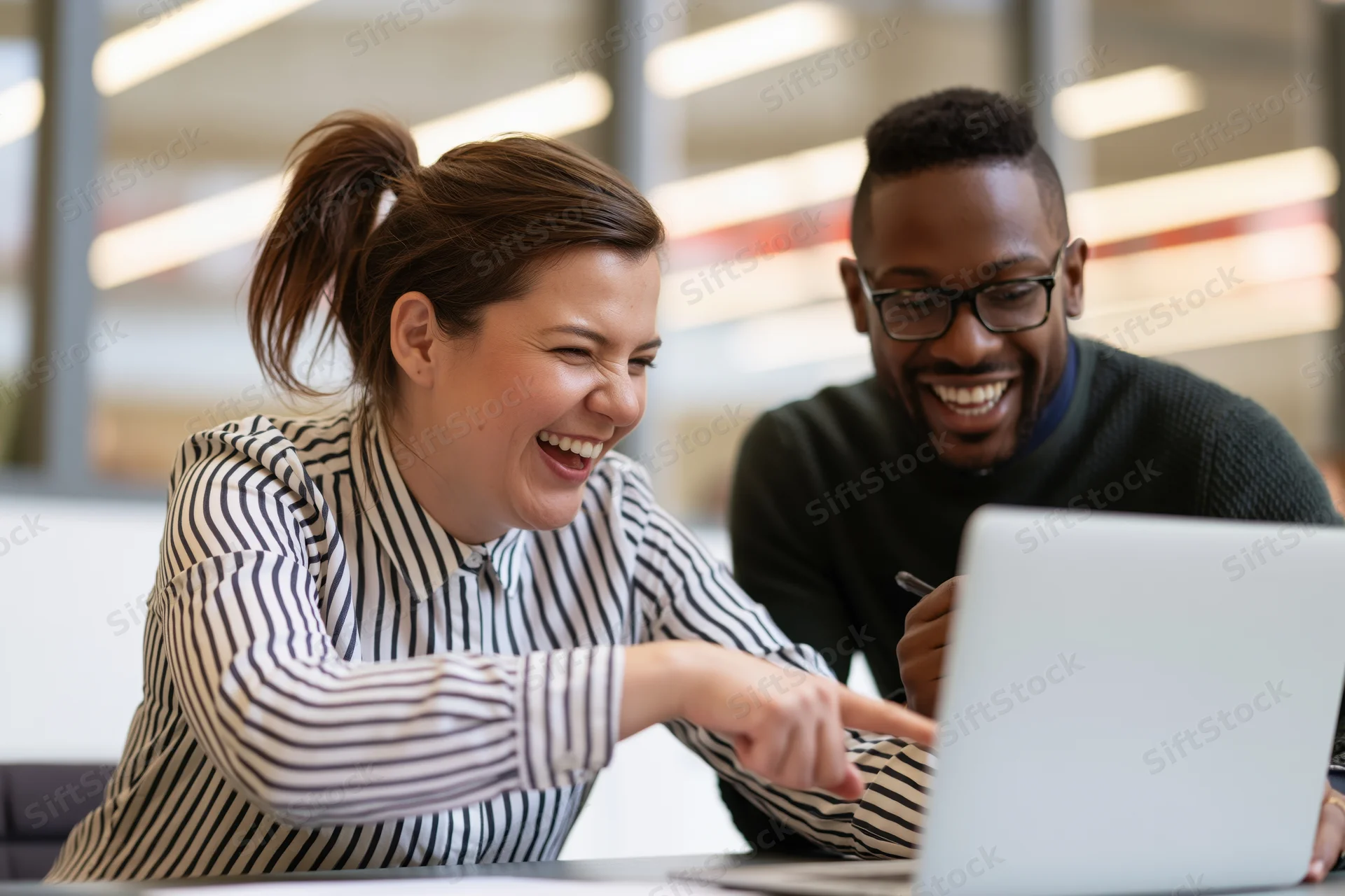 Two colleagues laughing and pointing at a laptop screen in an office setting