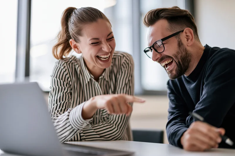 Two colleagues laughing and pointing at a laptop screen in an office meeting