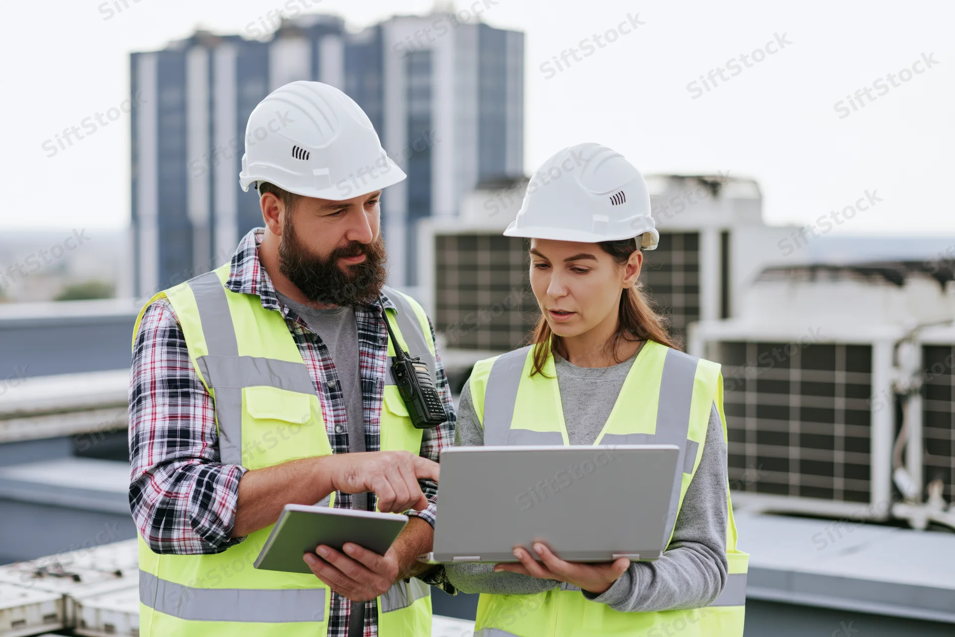 Two construction engineers reviewing data on a laptop and tablet on a rooftop site
