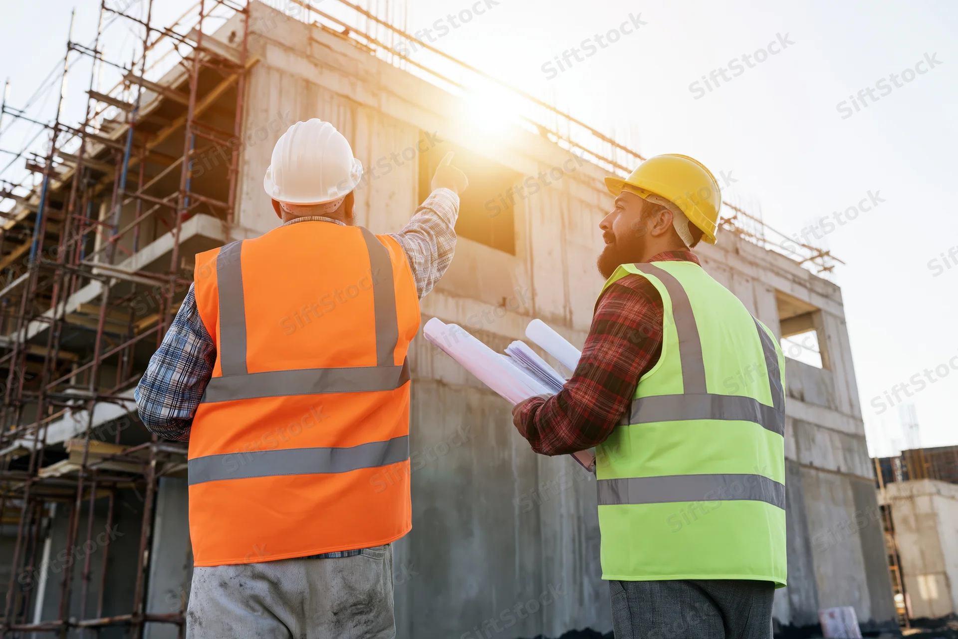 Two construction engineers discussing a building project on site, pointing at structure while holding blueprints