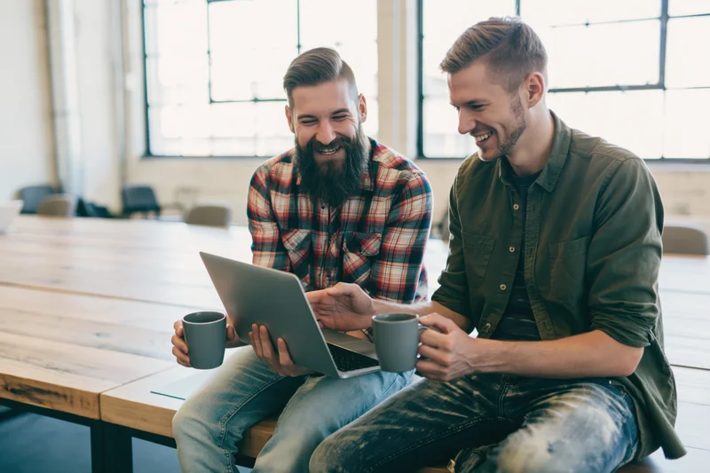 Two men laughing while using a laptop together at a wooden table in an office
