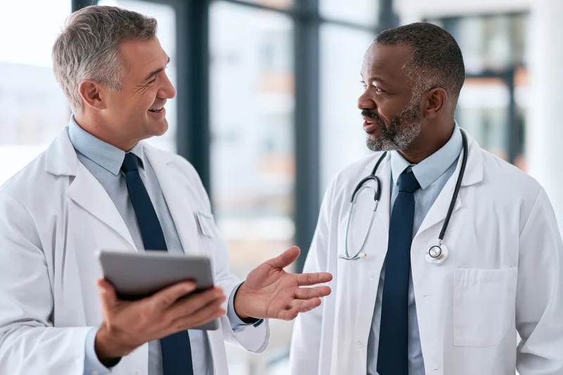 Two male doctors in conversation in a modern hospital office, one holding a tablet and gesturing while the other listens, both wearing lab coats and ties