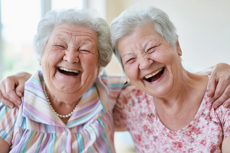 Two elderly women laughing together with arms around each other's shoulders indoors