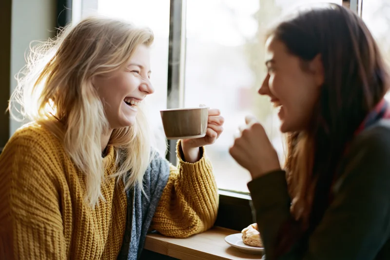 Two women laughing together at a cafe counter holding coffee cups