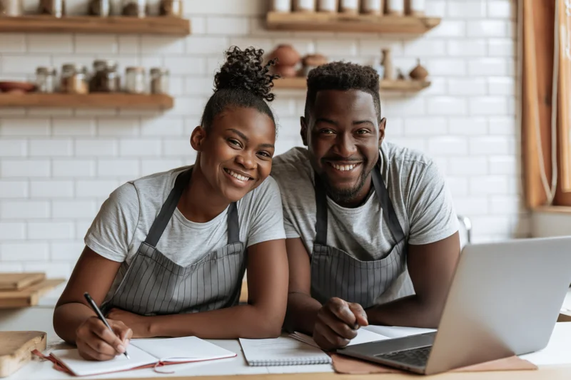 Young black couple in aprons smiling at camera while planning business at cafe counter with laptop and notebook
