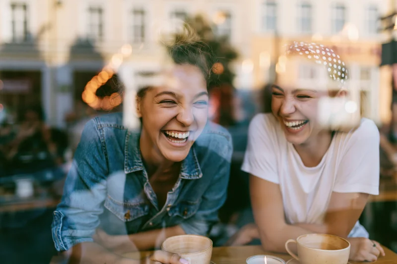 Two young women laughing together over coffee at a cafe viewed through a window