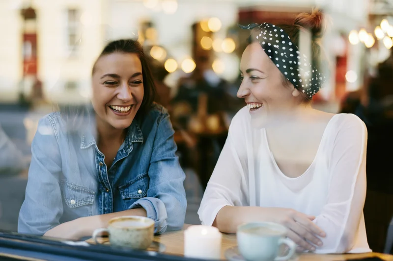 Two young women laughing together over coffee at a cafe table viewed through a window