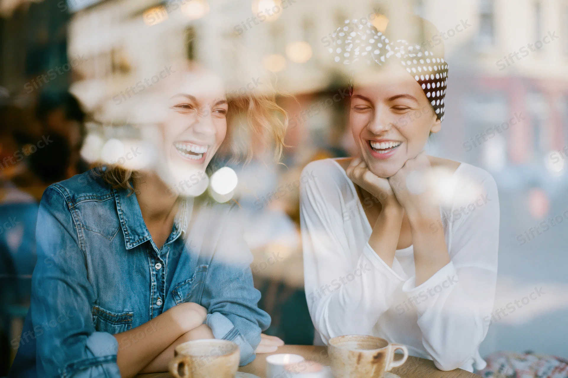 Two young women laughing together over coffee at a cafe viewed through a window with reflections