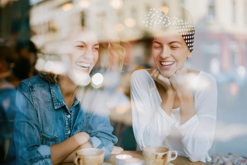 Two young women laughing together over coffee at a cafe viewed through a window with reflections