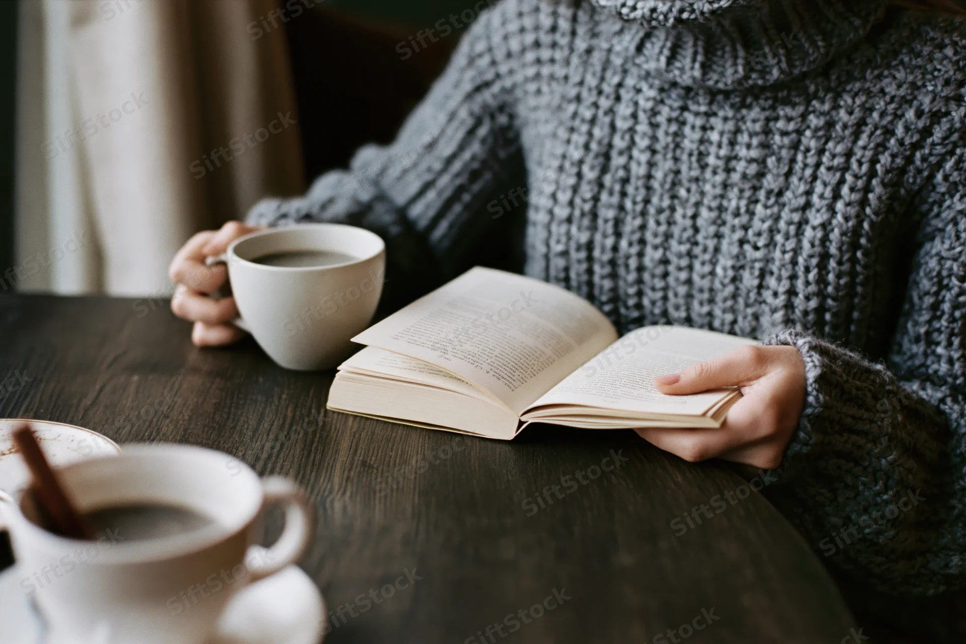 Woman reading a book while holding a coffee cup at a wooden table