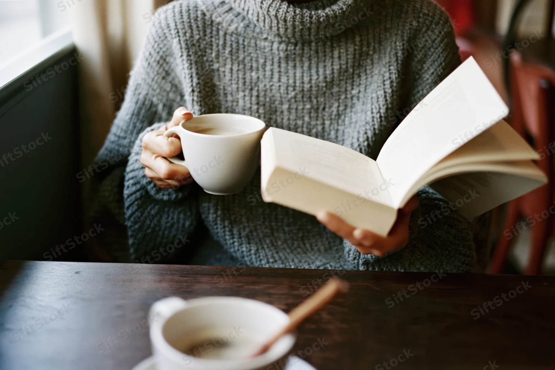 Woman reading a book while holding a white coffee cup at a wooden table near a window