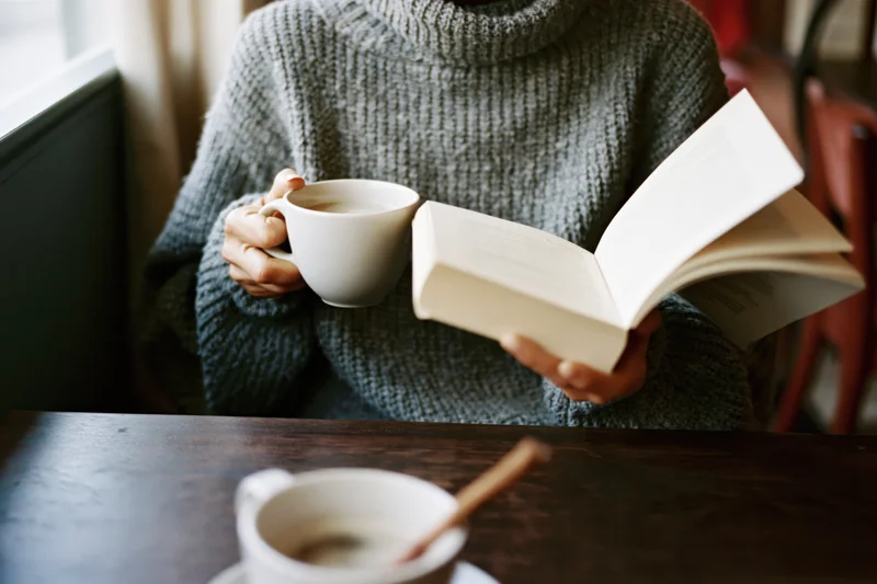 Woman reading a book while holding a white coffee cup at a wooden table near a window
