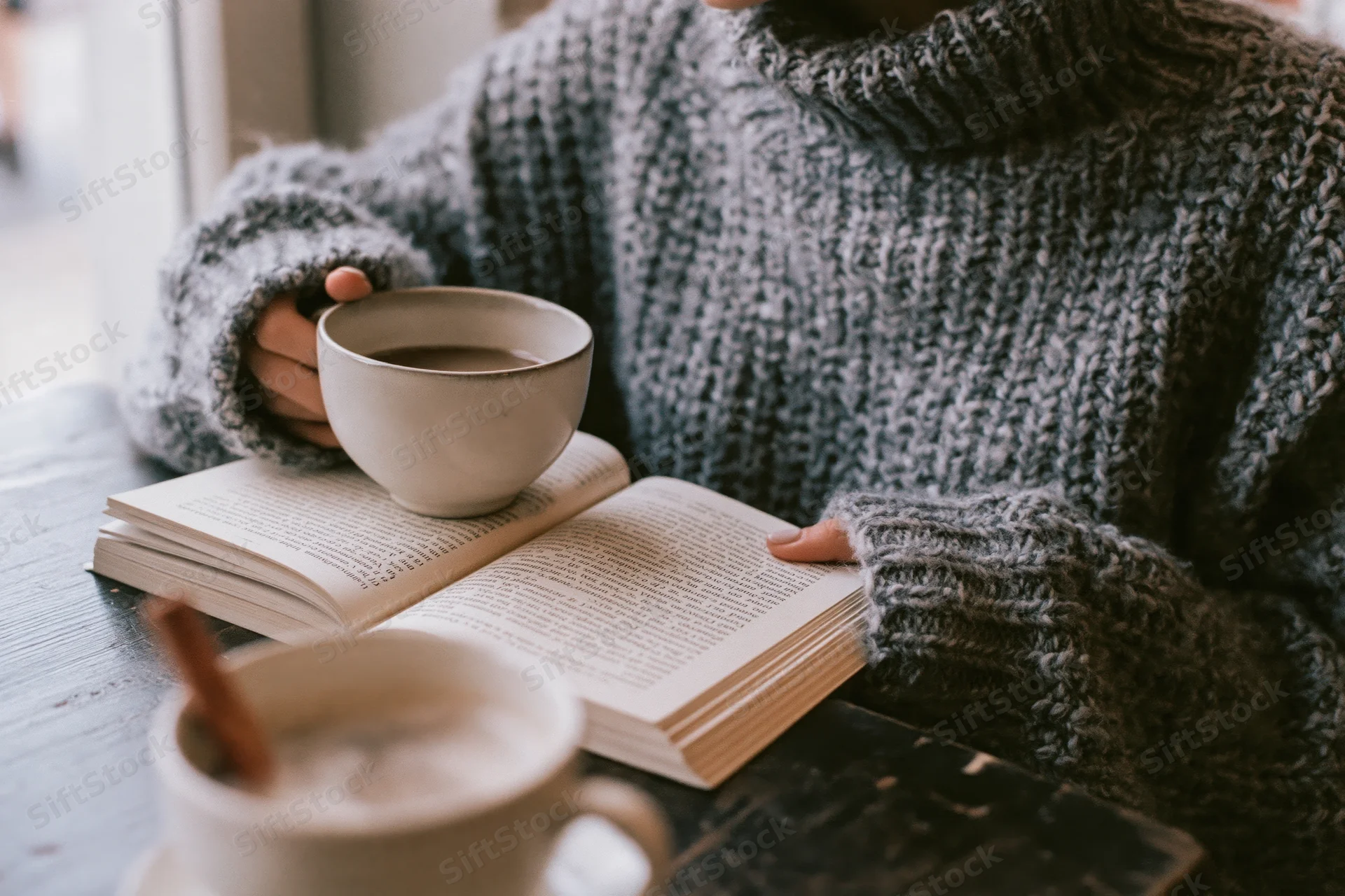 Woman in chunky grey turtleneck sweater reading a book while holding a white ceramic coffee cup on a wooden table