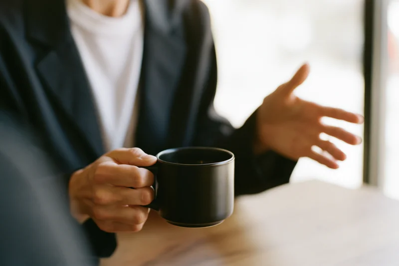 Woman in dark blazer holding black ceramic mug while gesturing during conversation at desk