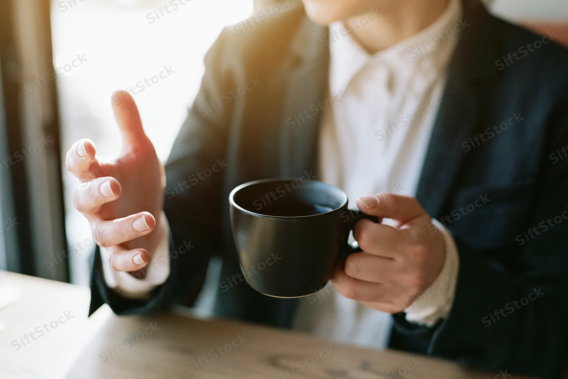 Woman in dark blazer and white shirt holding a black ceramic mug while gesturing during a meeting at a wooden table