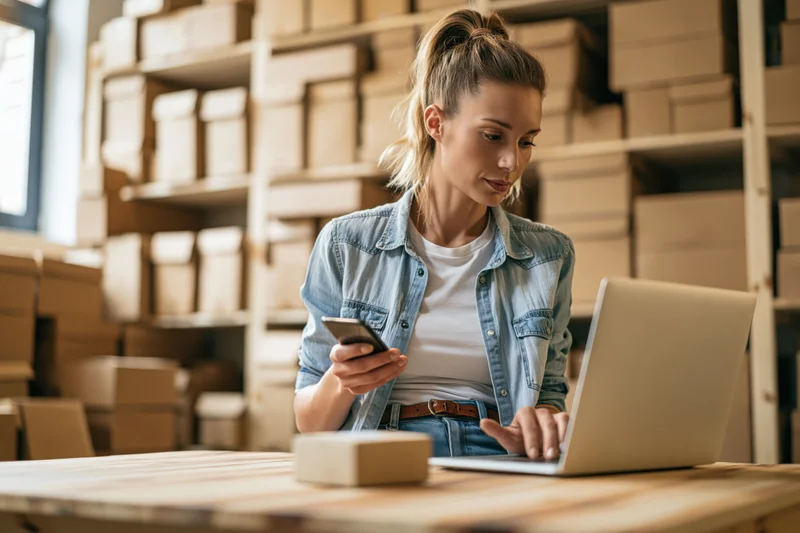 Woman using laptop and smartphone at desk in warehouse with boxes on shelves