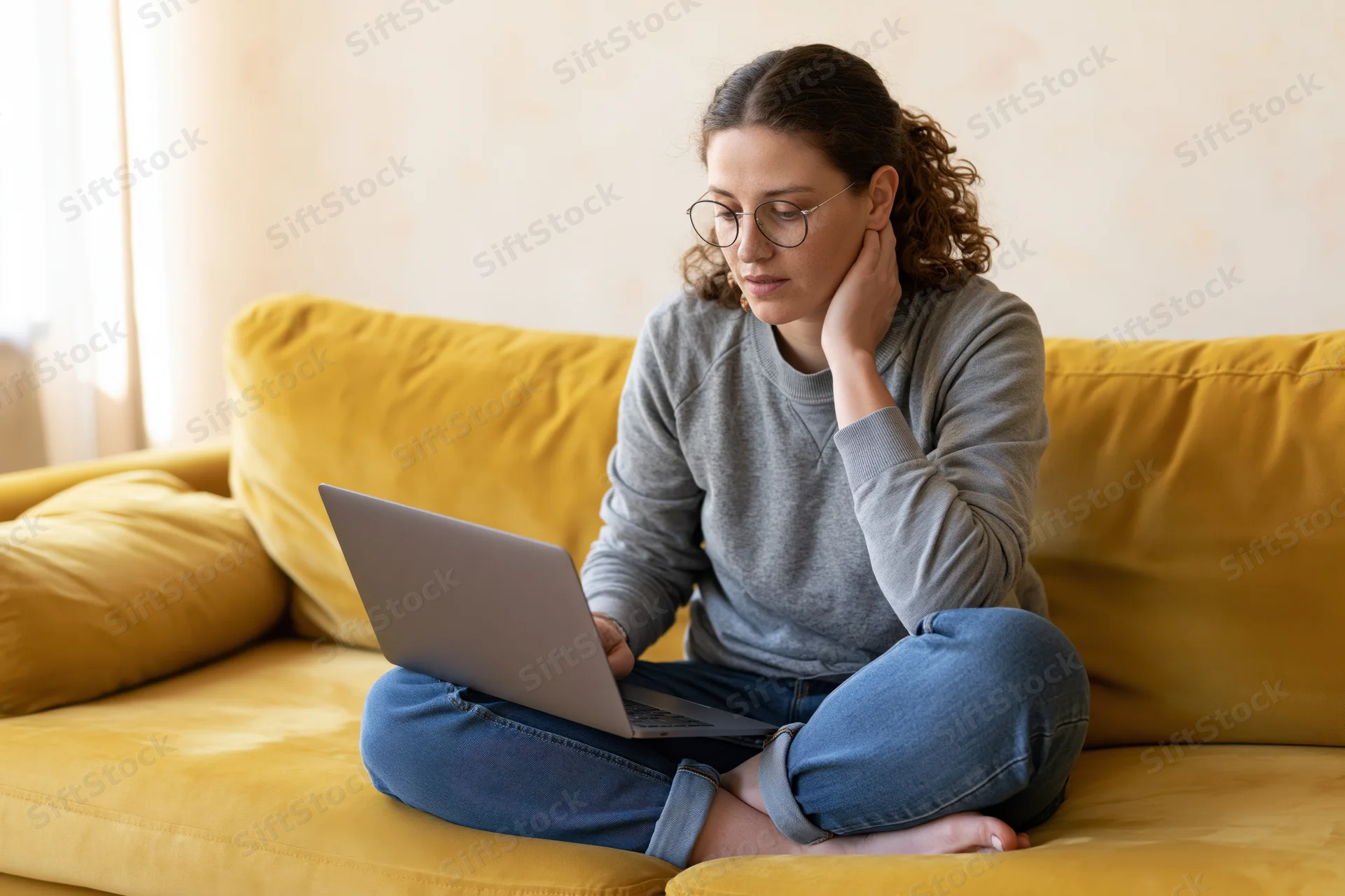 Woman sitting cross-legged on yellow sofa using laptop at home