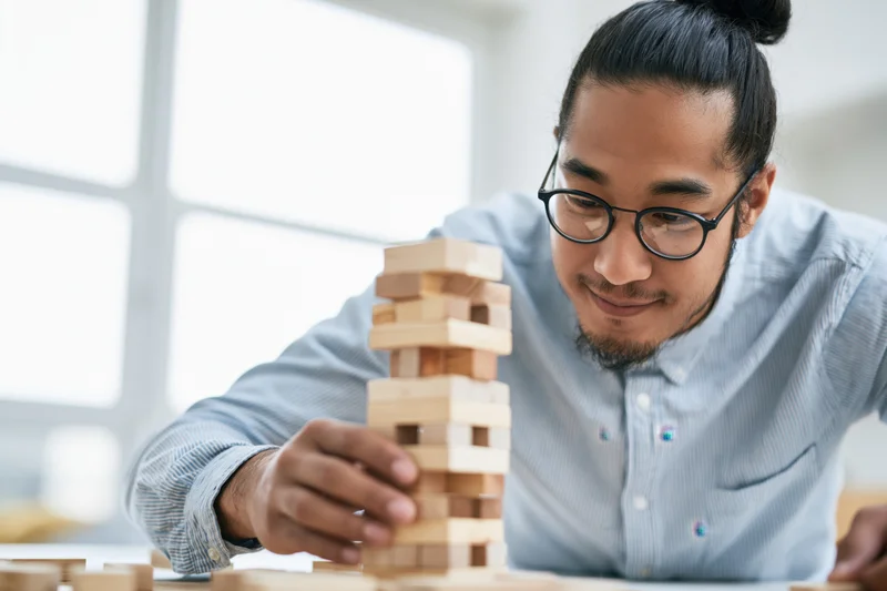 Young asian man with glasses and beard carefully stacking wooden blocks at a desk in a bright office