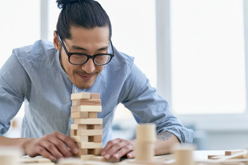 Young asian man with glasses and hair in a bun carefully stacking wooden blocks at a desk in an office setting