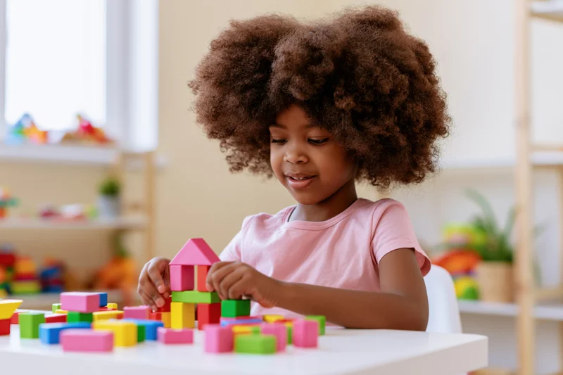 Young black girl smiling while building with colorful blocks at a white table in a playroom