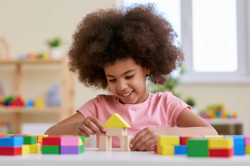Young black girl smiling while building a house with colorful wooden blocks at a table in a playroom
