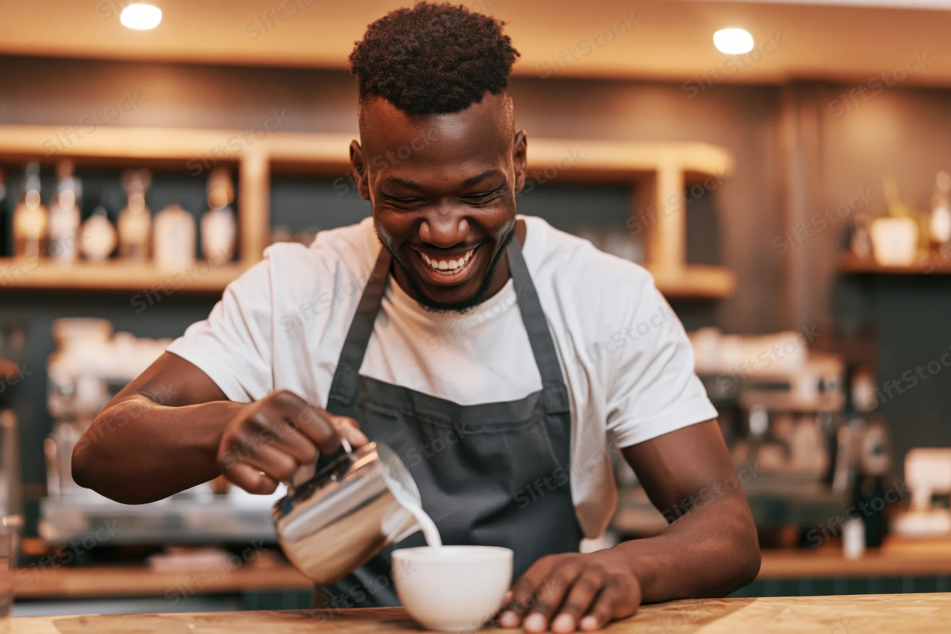 Young black male barista smiling while pouring steamed milk into a white cup at a cafe counter