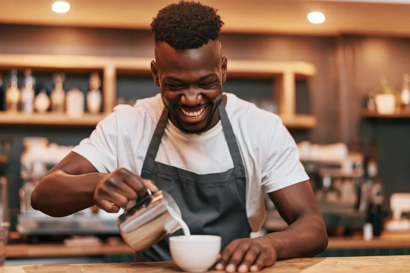 Young black male barista smiling while pouring steamed milk into a white cup at a cafe counter