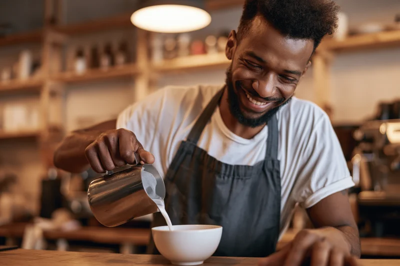 Smiling black male barista pouring steamed milk into a white cup at a cafe counter