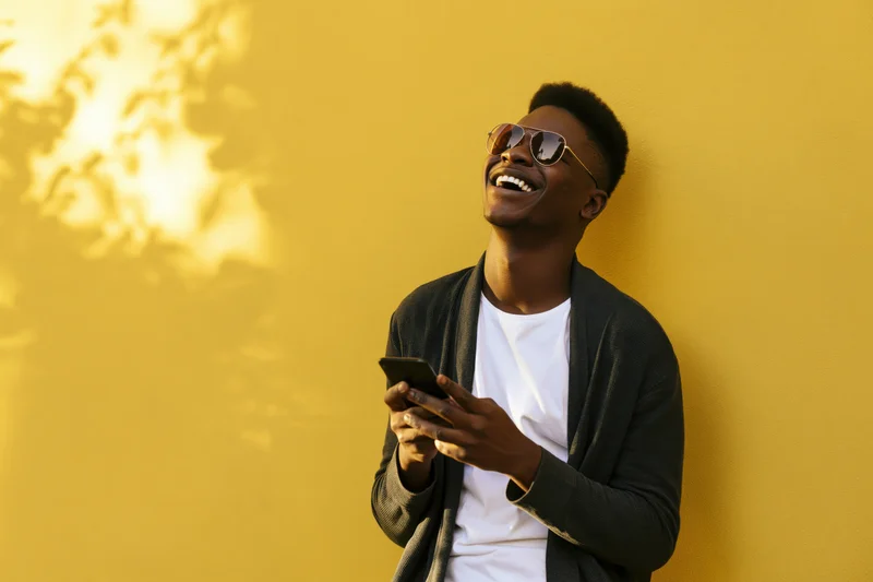 Young black man laughing while holding smartphone against yellow wall outdoors