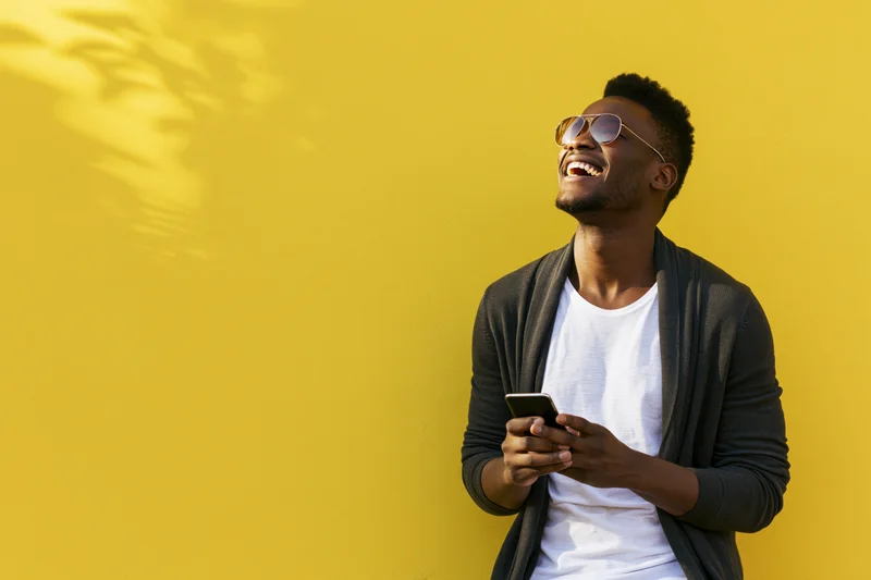 Young black man laughing while holding smartphone against yellow wall outdoors