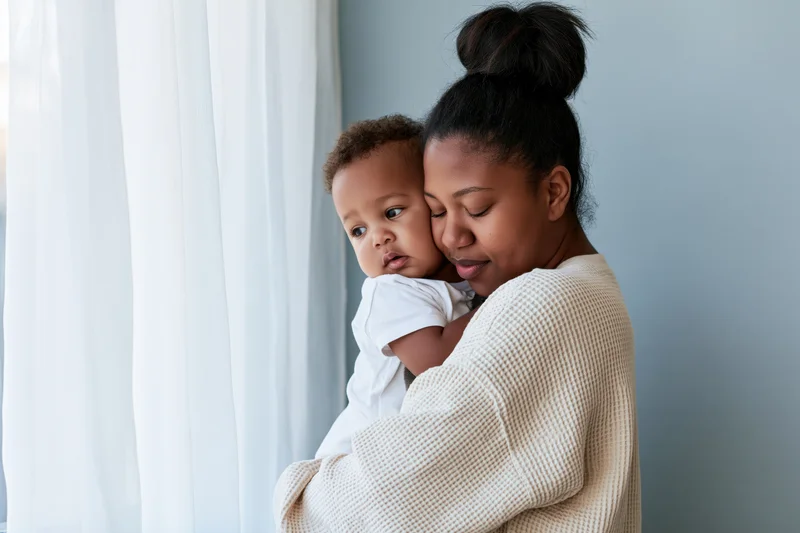 Black mother holding toddler son close by window with eyes closed