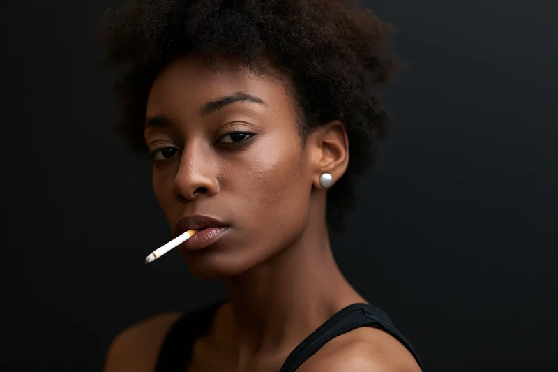 Young black woman holding unlit cigarette between lips in studio portrait with dark background