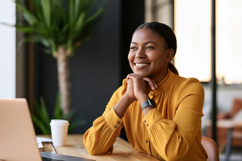 Smiling black woman sitting at wooden desk with clasped hands beside laptop in modern office