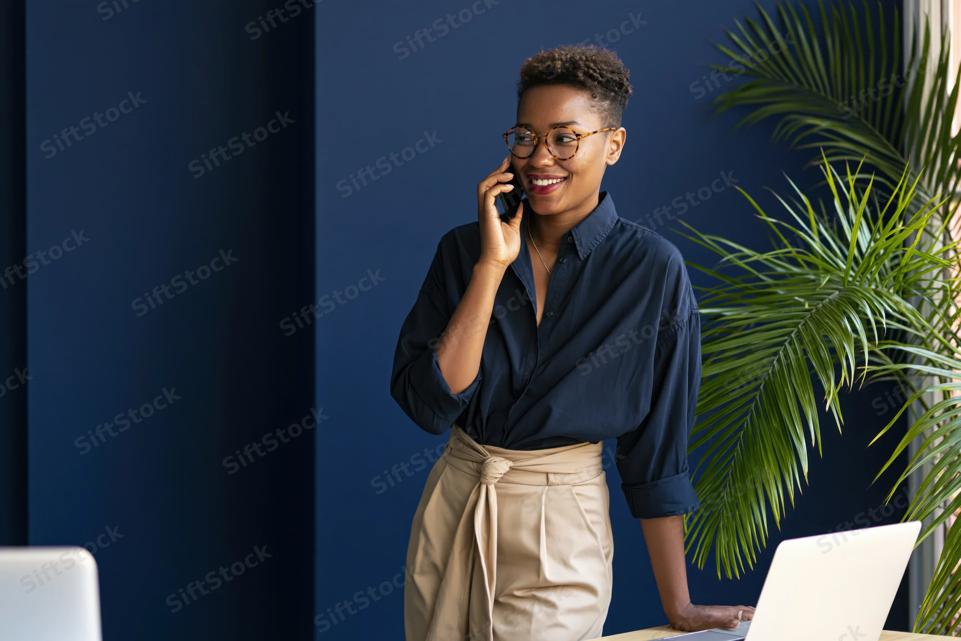 Young black woman smiling while talking on smartphone at office desk with laptop and plant nearby