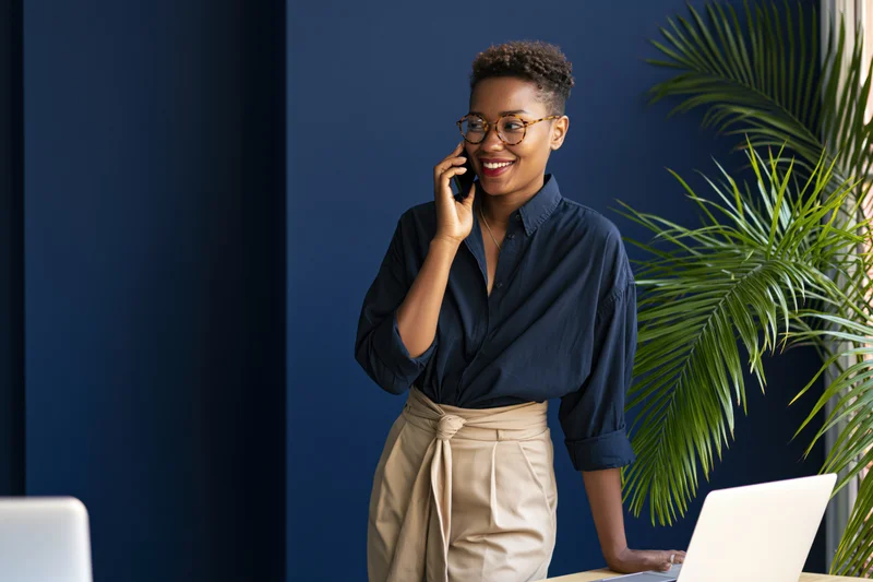 Young black woman smiling while talking on smartphone at office desk with laptop and plant nearby