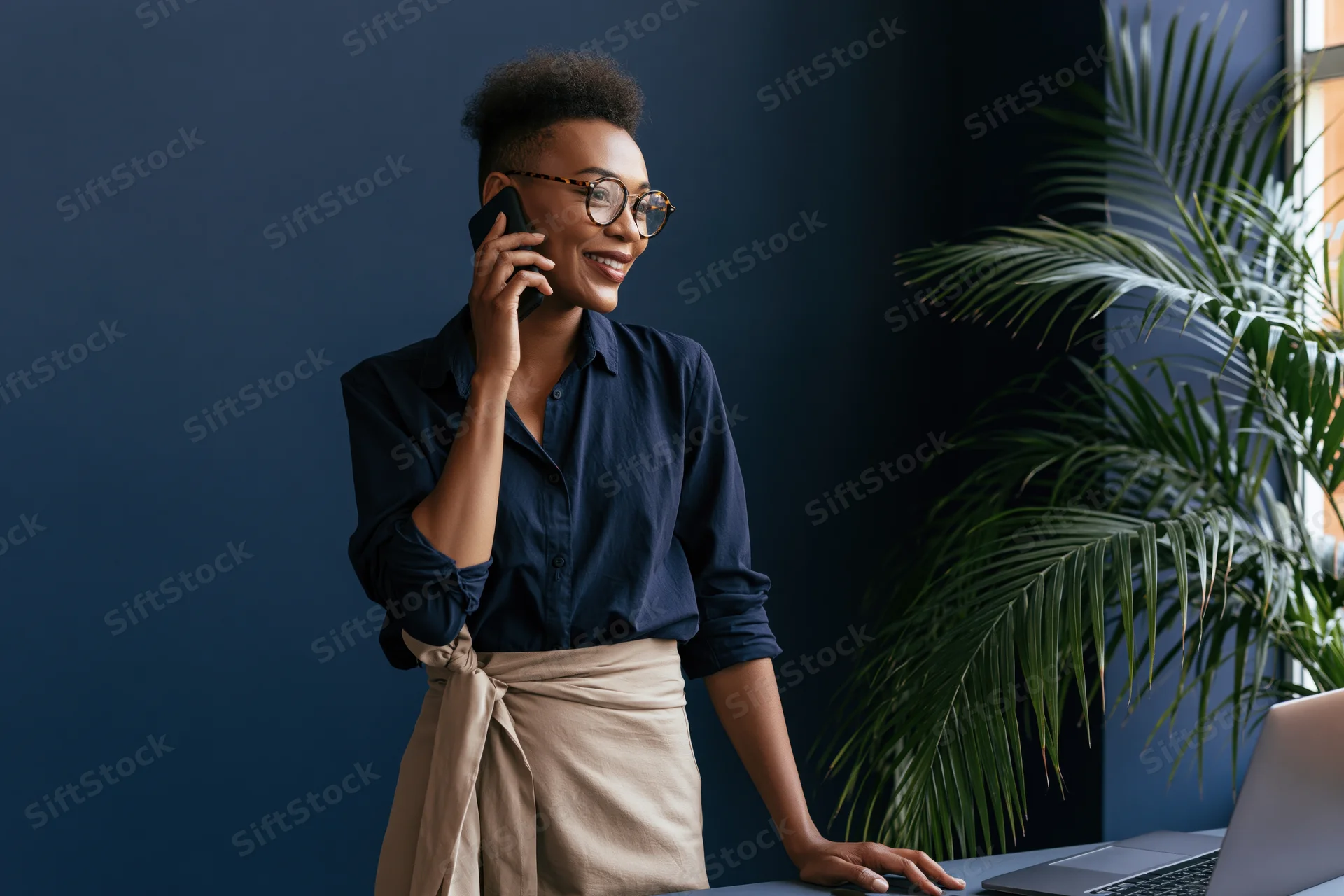 Black woman smiling while talking on smartphone at office desk with laptop and plant nearby