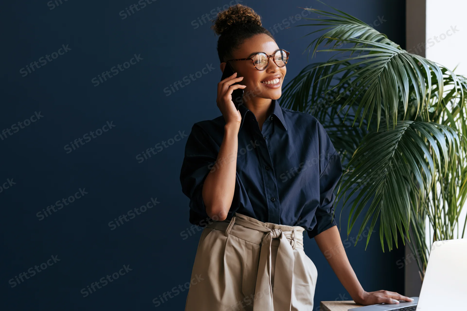 Young black woman smiling while talking on smartphone at desk with laptop and plant nearby