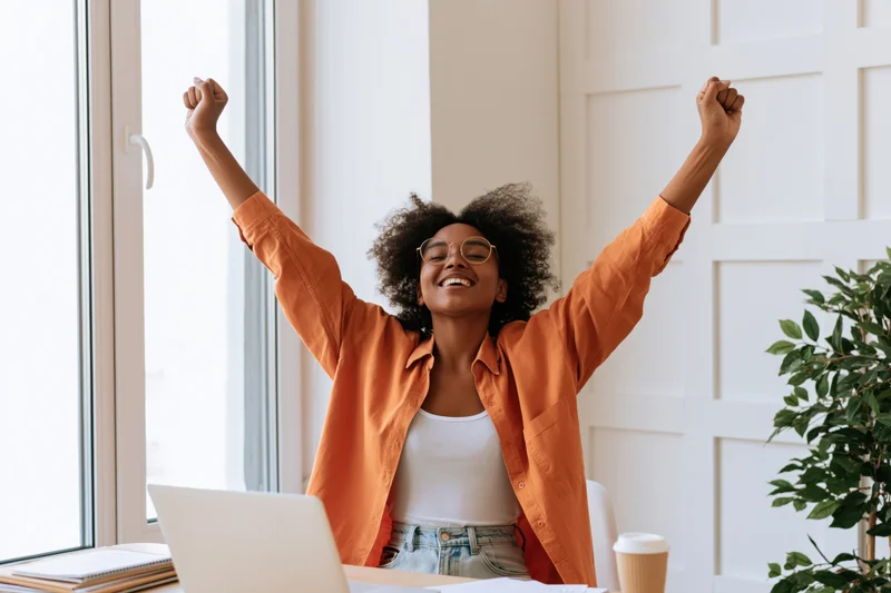Young black woman celebrating with arms raised at her desk in a bright home office