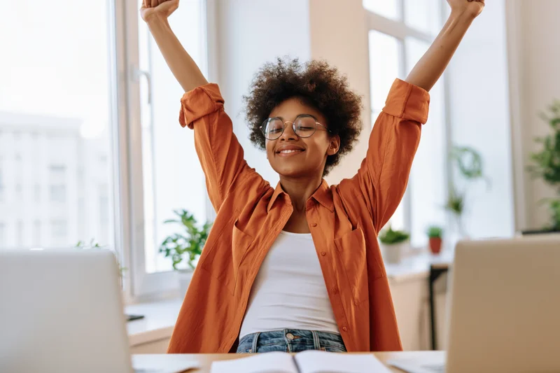 Young black woman celebrating with arms raised at office desk
