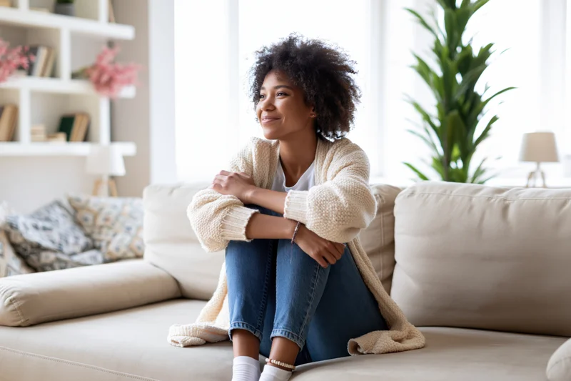 Young black woman sitting on beige sofa smiling with arms crossed in living room