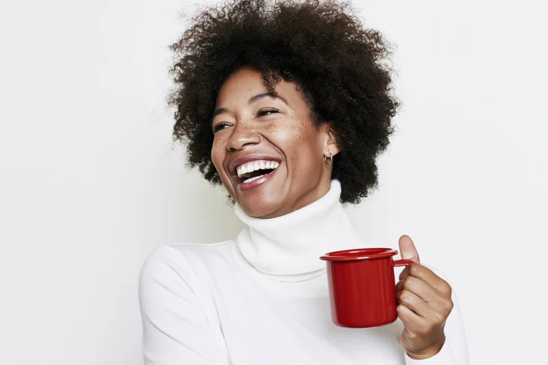 Young black woman laughing while holding a red enamel mug against a white background