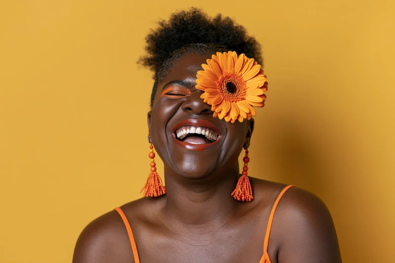Black woman laughing with orange flower over her eye against yellow background
