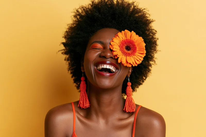 Black woman laughing with orange flower over eye against yellow background