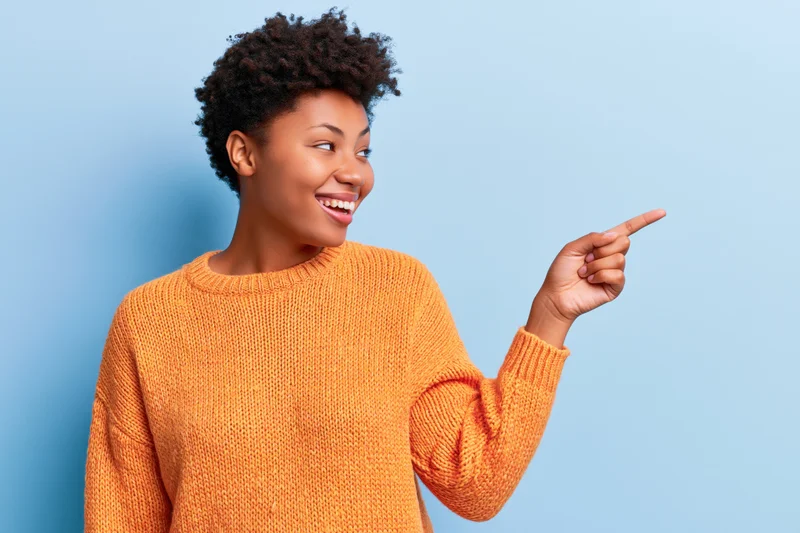 Young black woman smiling and pointing to the side in orange sweater against blue background