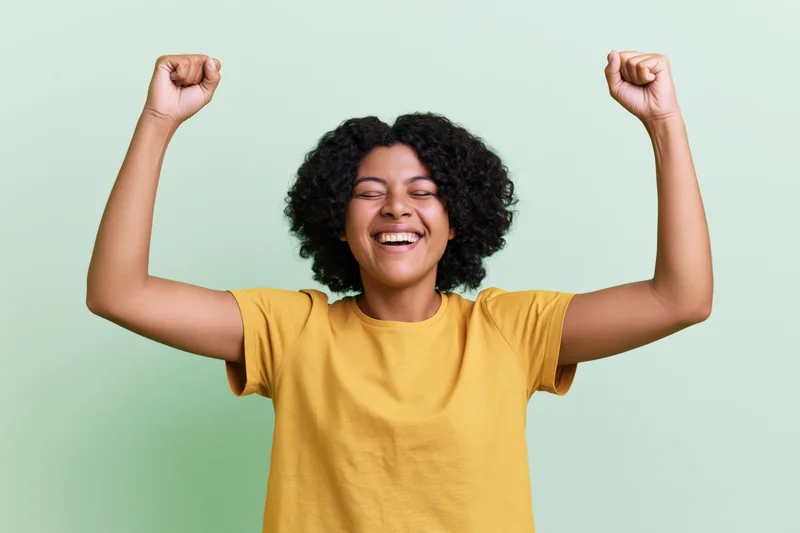 Young woman laughing with eyes closed and fists raised in celebration against a light green background